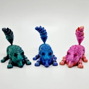 May include: Three 3D printed figurines of stylized foxes in different colors: teal, blue, and pink. Each figurine has a curled tail and a detailed face.