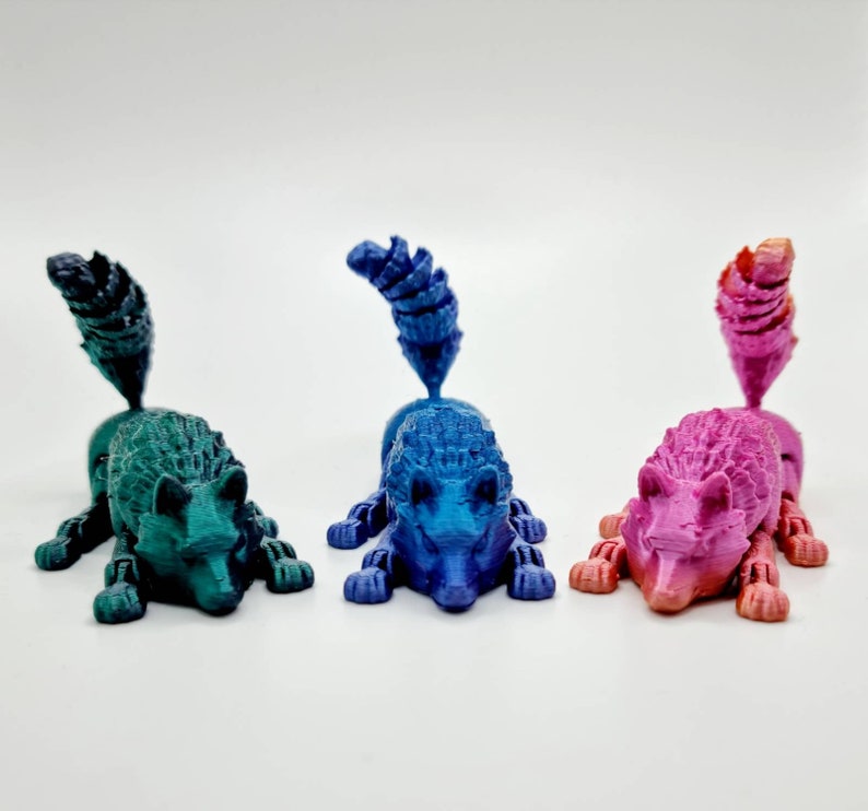 May include: Three 3D printed figurines of stylized foxes in different colors: teal, blue, and pink. Each figurine has a curled tail and a detailed face.