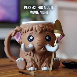 May include: A brown mammoth-shaped mug with a gold spoon. The mug has large black eyes, white tusks, and pink inner ears. The text "PERFECT FOR A CUTE MOVIE NIGHT" is above the mug.