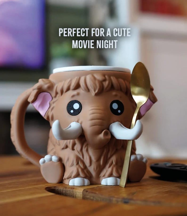 May include: A brown mammoth-shaped mug with a gold spoon. The mug has large black eyes, white tusks, and pink inner ears. The text "PERFECT FOR A CUTE MOVIE NIGHT" is above the mug.