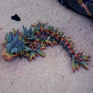 May include: A colorful, spiky, and detailed creature figurine made of plastic. The creature is green, orange, and red with a black eye. It is lying on a sandy surface.