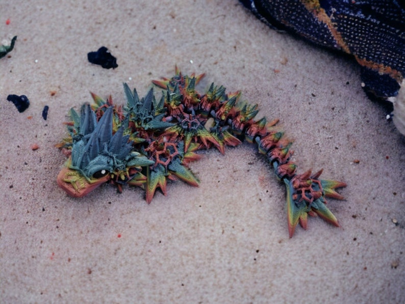 May include: A colorful, spiky, and detailed creature figurine made of plastic. The creature is green, orange, and red with a black eye. It is lying on a sandy surface.