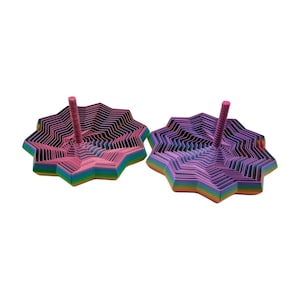 May include: Two colorful, star-shaped spinning tops. One is pink and the other is purple. Each has a central pink peg. The tops are made of layered, colorful paper, with rainbow edges. The background is white.