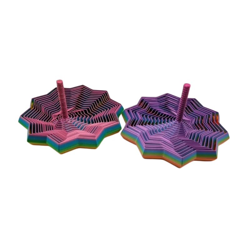 May include: Two colorful, star-shaped spinning tops. One is pink and the other is purple. Each has a central pink peg. The tops are made of layered, colorful paper, with rainbow edges. The background is white.