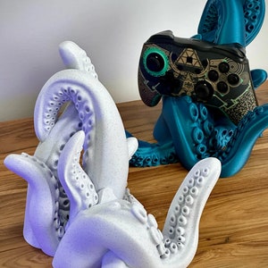 May include: Two 3D printed octopus-shaped game controller holders. One is teal blue and the other is white with a speckled finish. Both holders have multiple tentacles with suction cups.