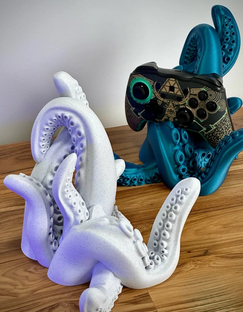May include: Two 3D printed octopus-shaped game controller holders. One is teal blue and the other is white with a speckled finish. Both holders have multiple tentacles with suction cups.