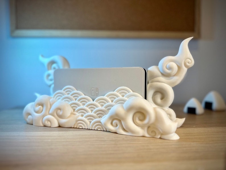 May include: White 3D printed Nintendo Switch stand with a cloud design. The stand has a wave pattern on the front and two cloud-shaped supports on the sides.