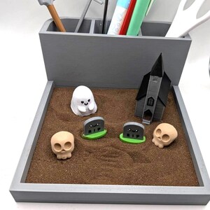 Pu&ograve; includere: Un organizzatore da scrivania in legno grigio con un piccolo vassoio di sabbia che presenta un fantasma bianco, due lapidi grigie con erba verde, una chiesa grigia e due teschi marroni. Il vassoio di sabbia &egrave; un organizzatore da scrivania a tema Halloween.