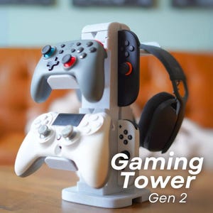 May include: A grey and white gaming tower holding two game controllers and a headset. The controllers are grey and white with red and blue buttons. The text "Gaming Tower Gen 2" is displayed on the front.
