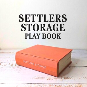 May include: An orange storage box shaped like a book, with the words "SETTLERS STORAGE PLAY BOOK" in black above it. The box has the words "SETTLERS STORAGE" printed on the spine. The box is sitting on a white and light brown wooden surface.