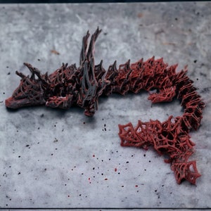 May include: An abstract sculpture resembling a dragon or serpent, predominantly red with dark grey detailing. The intricate, openwork design is set against a mottled grey surface. The artwork is a unique, decorative piece.