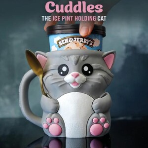 May include: A gray cat-shaped mug with pink accents, holding a pint of ice cream and a golden spoon. The mug has large black eyes and the text "Cuddles" and "THE ICE PINT HOLDING CAT" in pink.