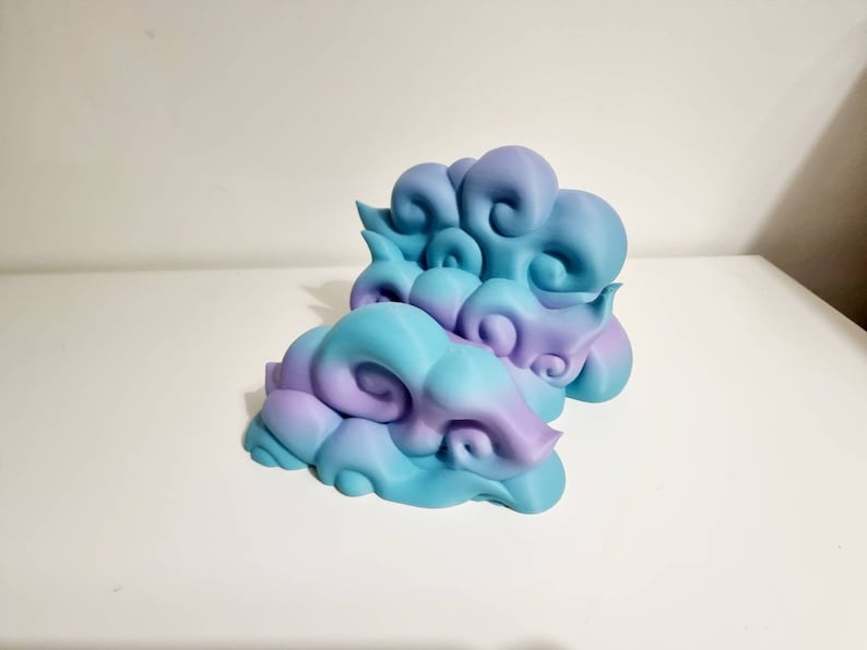 May include: Two decorative cloud sculptures in shades of blue and purple. The clouds have a swirling, textured design and are placed on a white surface. The sculptures are a unique home decor item.