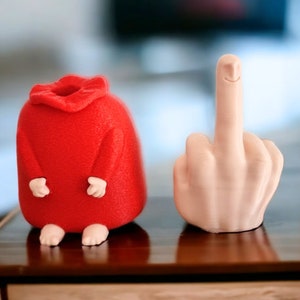 May include: A red felt figurine with a smiling face and a white ceramic figurine of a hand giving the middle finger.