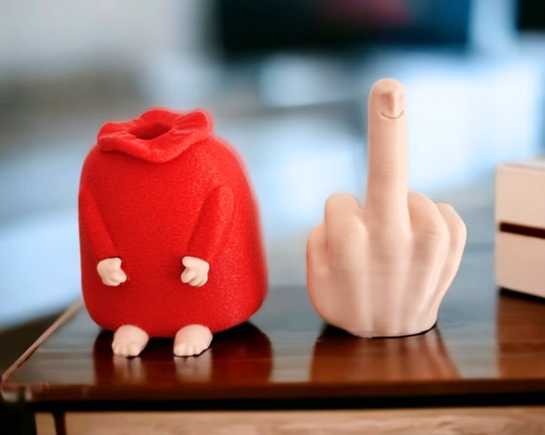 May include: A red felt figurine with a smiling face and a white ceramic figurine of a hand giving the middle finger.