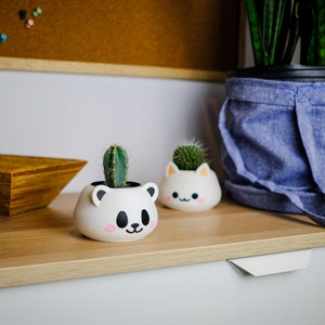 May include: Two white ceramic planters shaped like animal faces, one a bear and one a cat, with small green cactus plants inside. The planters are sitting on a light wood surface with a wooden bowl and a blue fabric pot with a green plant in the background.