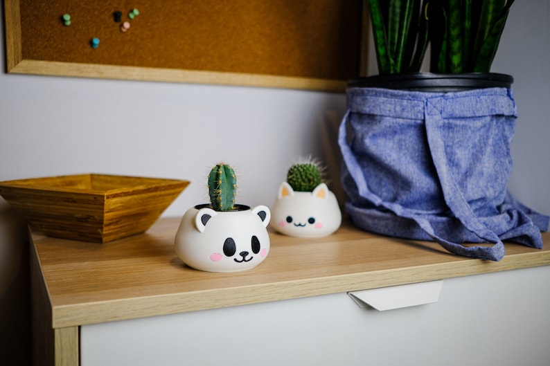 May include: Two white ceramic planters shaped like animal faces, one a bear and one a cat, with small green cactus plants inside. The planters are sitting on a light wood surface with a wooden bowl and a blue fabric pot with a green plant in the background.