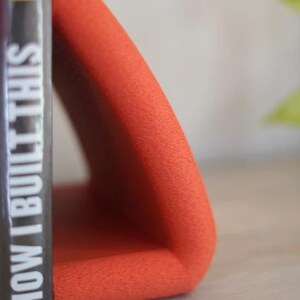 May include: An orange, triangular bookend with a textured surface. A book with the title "HOW I BUILT THIS" is placed next to the bookend. The bookend is made of a soft material and is designed to hold books upright.