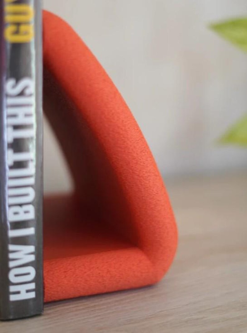May include: An orange, triangular bookend with a textured surface. A book with the title "HOW I BUILT THIS" is placed next to the bookend. The bookend is made of a soft material and is designed to hold books upright.