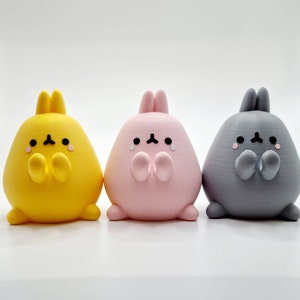 K&ouml;nnte beinhalten: Drei kleine Pl&uuml;schtiere in Form von Hasen mit unterschiedlichem Fell. Der Hase links ist gelb, der mittlere Hase ist rosa und der Hase rechts ist grau. Jeder Hase hat eine kleine, schwarze Nase und Augen sowie einen kleinen, schwarzen Mund.