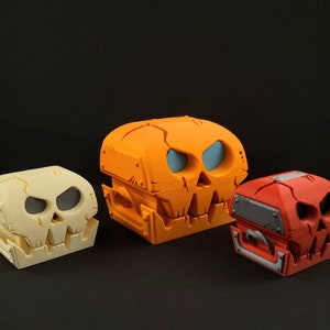 May include: Three 3D printed skull-shaped boxes. The largest box is orange, the middle box is white, and the smallest box is red.
