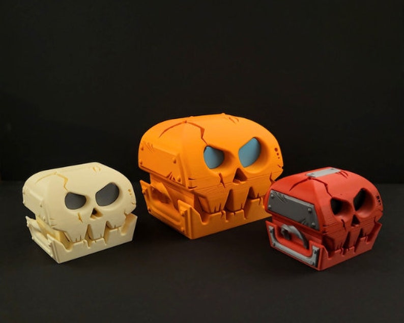May include: Three 3D printed skull-shaped boxes. The largest box is orange, the middle box is white, and the smallest box is red.