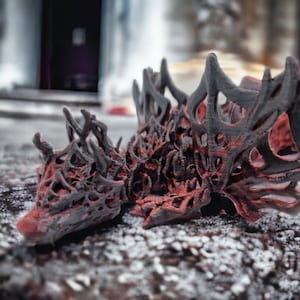 May include: A detailed, 3D-printed dragon sculpture with an intricate, openwork design. The dragon is mainly dark grey with red accents, and rests on a textured, stone-like surface. The sculpture's intricate details are highlighted by the contrast of colours.