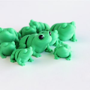 May include: A collection of green frog figurines with black eyes. The frogs vary in size and are clustered together, creating a playful and whimsical scene. The figurines are made of a smooth material and are set against a white background.