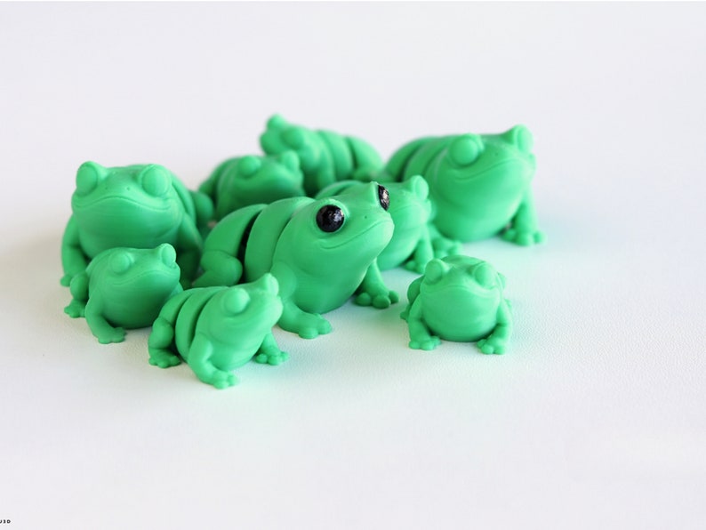 May include: A collection of green frog figurines with black eyes. The frogs vary in size and are clustered together, creating a playful and whimsical scene. The figurines are made of a smooth material and are set against a white background.