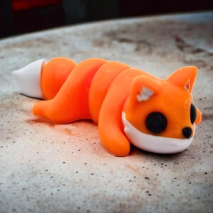 May include: Orange and white curled fox figurine with black eyes. The figurine is lying on a light brown surface.
