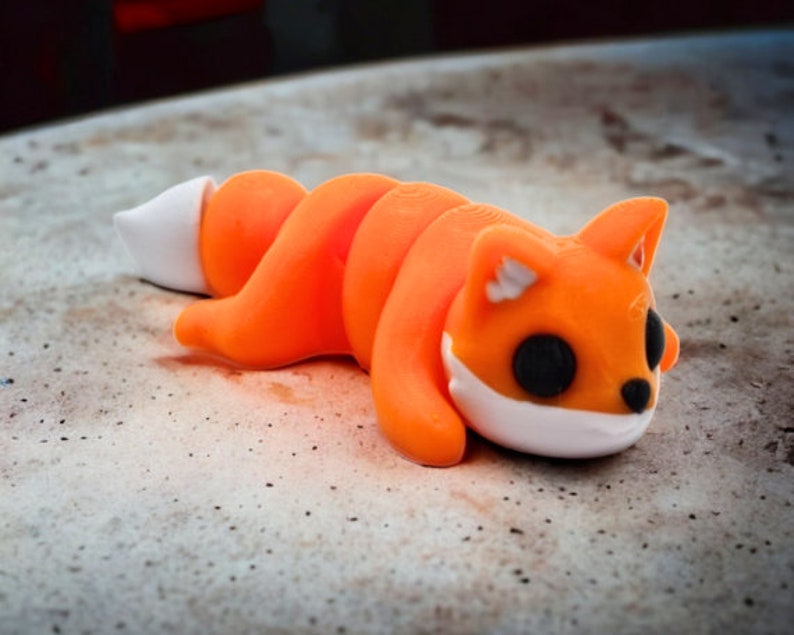 May include: Orange and white curled fox figurine with black eyes. The figurine is lying on a light brown surface.