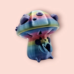 May include: A rainbow-colored mushroom figurine with a whimsical design. The mushroom has a wide, flat cap with a textured surface and a small, round stem. The cap is decorated with purple dots and the stem has a blue and purple swirl pattern.