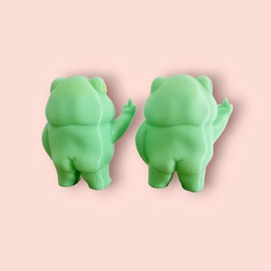 May include: Two green frog figurines with yellow eyes, both giving the middle finger. The frogs are standing on a pink surface.