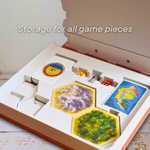 May include: Open game box with compartments for game pieces. The box contains cards, dice, and hexagonal tiles with landscape illustrations. The text "Storage for all game pieces" is visible on the top.