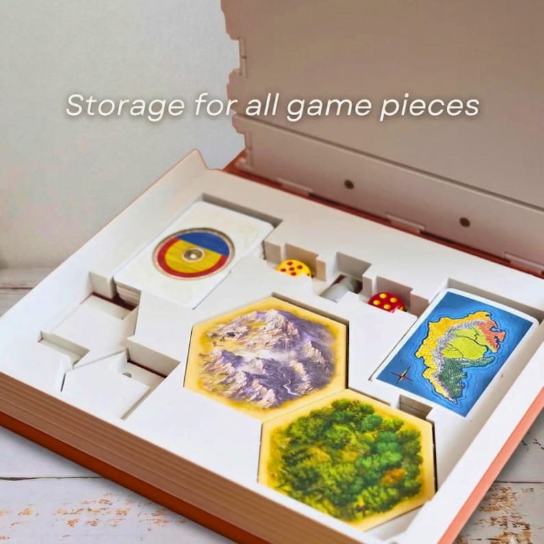 May include: Open game box with compartments for game pieces. The box contains cards, dice, and hexagonal tiles with landscape illustrations. The text "Storage for all game pieces" is visible on the top.