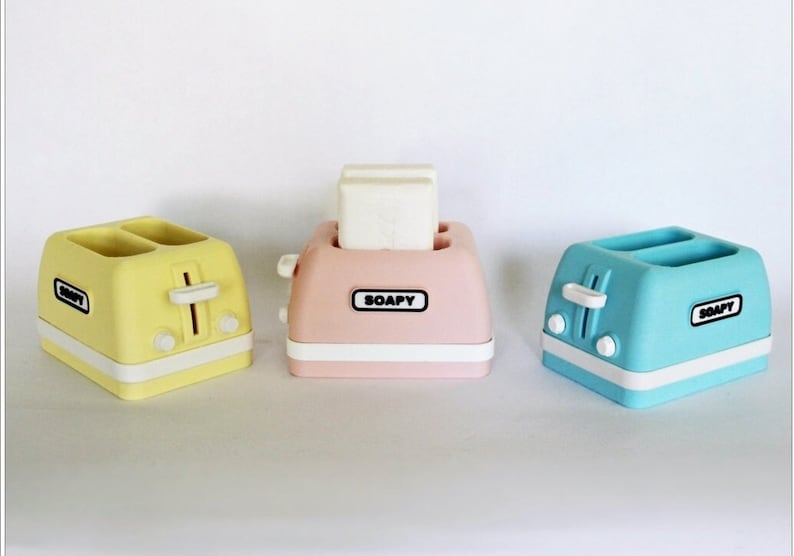 K&ouml;nnte beinhalten: Drei Miniatur-Toaster in Gelb, Rosa und Blau mit dem Wort "SOAPY" auf der Vorderseite. Der rosafarbene Toaster enth&auml;lt einen wei&szlig;en Seifenblock.