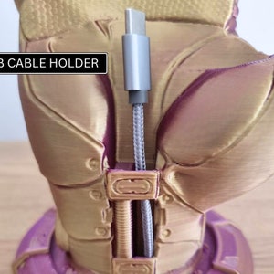 May include: A gold-colored 3D printed hand with a purple base, holding a gray USB cable. The hand has a textured surface and the text "USB CABLE HOLDER" is printed on a black rectangle.