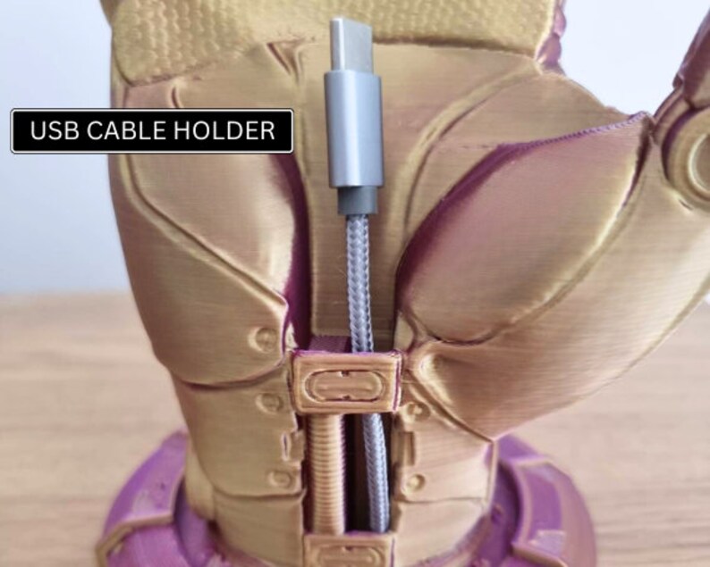 May include: A gold-colored 3D printed hand with a purple base, holding a gray USB cable. The hand has a textured surface and the text "USB CABLE HOLDER" is printed on a black rectangle.