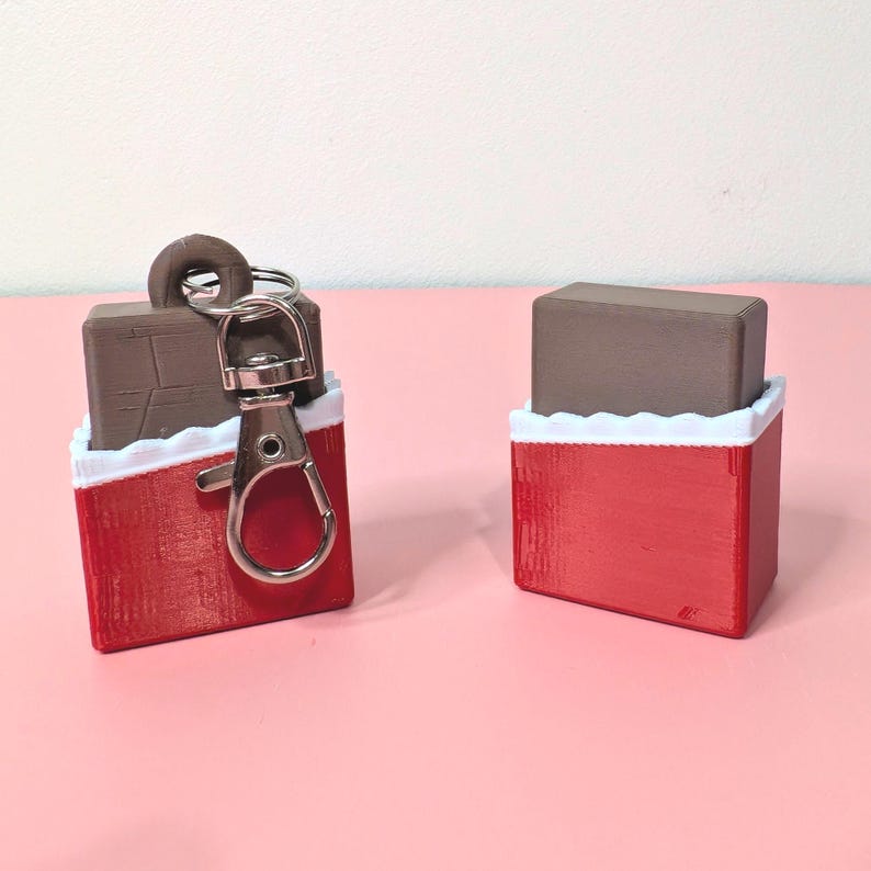 Puede incluir: Dos llaveros con forma de caja de regalo roja y blanca con un objeto rectangular marr&oacute;n en el interior. Un llavero tiene un mosquet&oacute;n y un llavero plateados. El otro llavero es una caja de regalo independiente. El fondo es rosa claro.