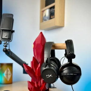 May include: Two black headphones with a red crystal-shaped stand. The headphones are on a wooden stand. The headphones on the left have a logo that says "HyperX". The headphones on the right have the text "DT 770 PRO" on the side.