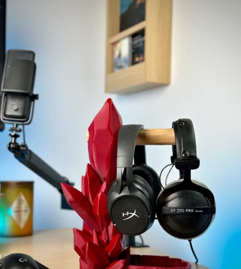 May include: Two black headphones with a red crystal-shaped stand. The headphones are on a wooden stand. The headphones on the left have a logo that says "HyperX". The headphones on the right have the text "DT 770 PRO" on the side.