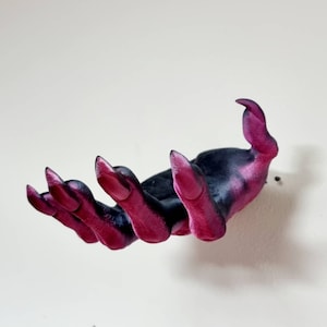 Puede incluir: Una mano impresa en 3D con largas y afiladas garras rosas. La mano es negra con detalles rosas y est&aacute; posada como si estuviera tendiendo la mano.