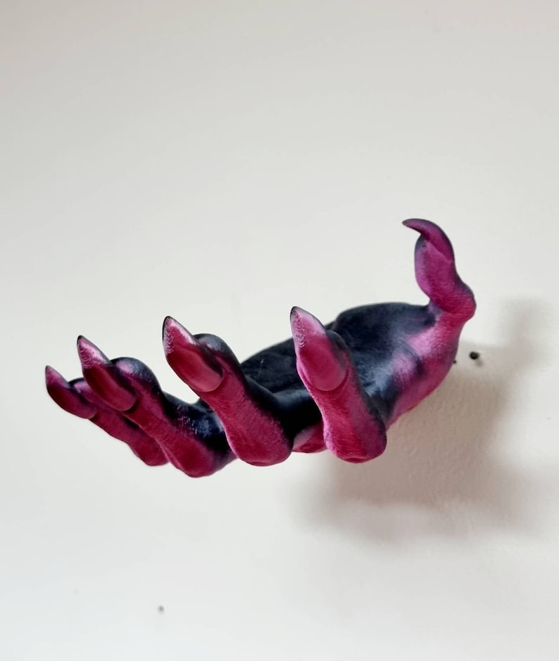 Puede incluir: Una mano impresa en 3D con largas y afiladas garras rosas. La mano es negra con detalles rosas y est&aacute; posada como si estuviera tendiendo la mano.