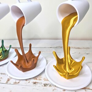 May include: White ceramic mugs appear to be pouring liquid into splash-shaped sculptures. One sculpture is brown, the other is gold, both resting on white saucers. The mugs are white with a ribbed texture. The background is a light wood.