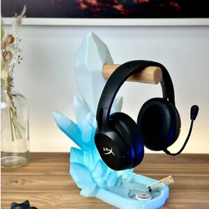 May include: A blue crystal-shaped headset stand with a wooden headset holder. The stand has a blue and white color scheme and is designed to hold a headset with a microphone. The stand is on a wooden surface.