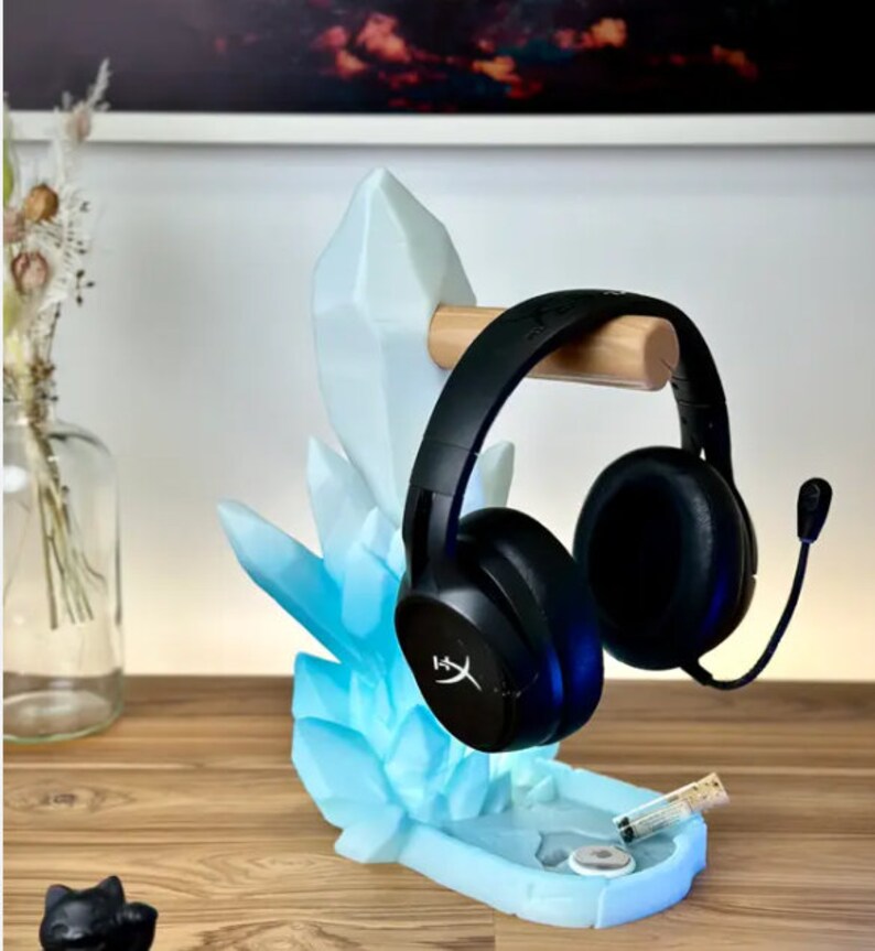 May include: A blue crystal-shaped headset stand with a wooden headset holder. The stand has a blue and white color scheme and is designed to hold a headset with a microphone. The stand is on a wooden surface.