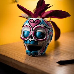 May include: A white, ceramic skull-shaped planter with blue, pink, and teal decorative accents. The planter holds a plant with deep burgundy leaves. The skull sits on a wooden surface against a yellow background.