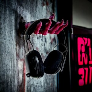 Puede incluir: Auriculares negros de diadema con un soporte en forma de mano rosa. Los auriculares tienen un dise&ntilde;o negro y plateado y est&aacute;n colgados en una pared.