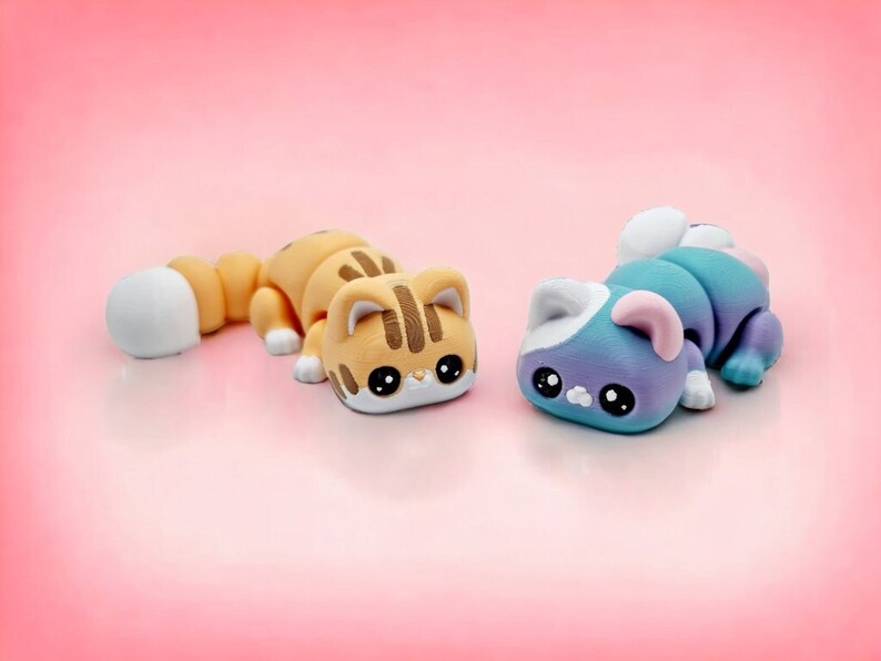 May include: Two small, hand-crafted cat figurines. One is light brown with white and brown stripes and a white tail. The other is light blue with a white tail and pink ears.