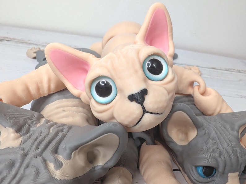 May include: A close-up of several Sphynx cat sculptures. The central cat has a light peach-colored body, large blue eyes, and pink-lined ears. Other cats are gray. The sculptures are detailed and appear to be made of a soft material.